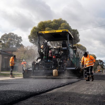Road construction crew operating a asphalt paver and smoothing fresh asphalt on a residential street.