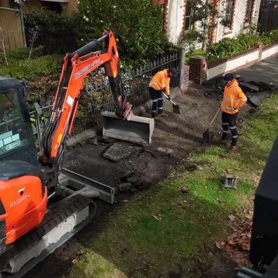 Construction workers using a mini excavator to remove and repair a residential footpath beside a house